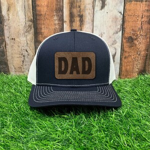 May include: A black and white trucker hat with a brown leather patch that says "DAD" on it.