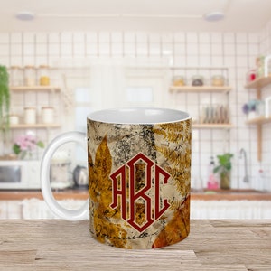 Puede incluir: Taza de cerámica blanca con un diseño marrón, amarillo y rojo con un monograma de las letras "AKC" y un patrón de hojas de otoño.