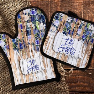May include: A set of two oven mitts with a rustic woodgrain background and blue floral accents. The mitts feature the text "The Smiths" in a blue script font.