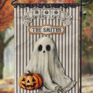 May include: Halloween garden flag featuring a ghost and jack-o'-lantern on a striped background. The flag reads "THE SMITHS" and has a decorative banner at the top. The flag is displayed on a black metal stand.