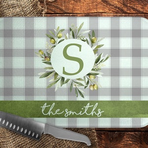 May include: A rectangular cutting board with a gray and white plaid pattern. The board features a green band with the text "the smiths" and a circular design with the letter "S" surrounded by olive branches. A stainless steel knife rests on the burlap.