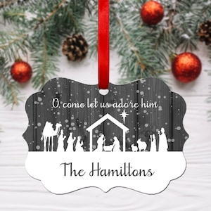 May include: A white metal Christmas ornament with a woodgrain background. The ornament features a silhouette of the nativity scene with the text "O come let us adore him" and "The Hamiltons".