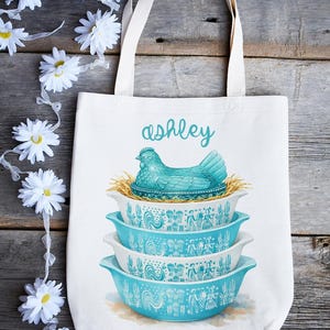 May include: White tote bag featuring a stack of three turquoise and white bowls with a rooster and floral design. A turquoise hen sits atop the bowls, with the name "ashley" in script above. The bag has white handles.