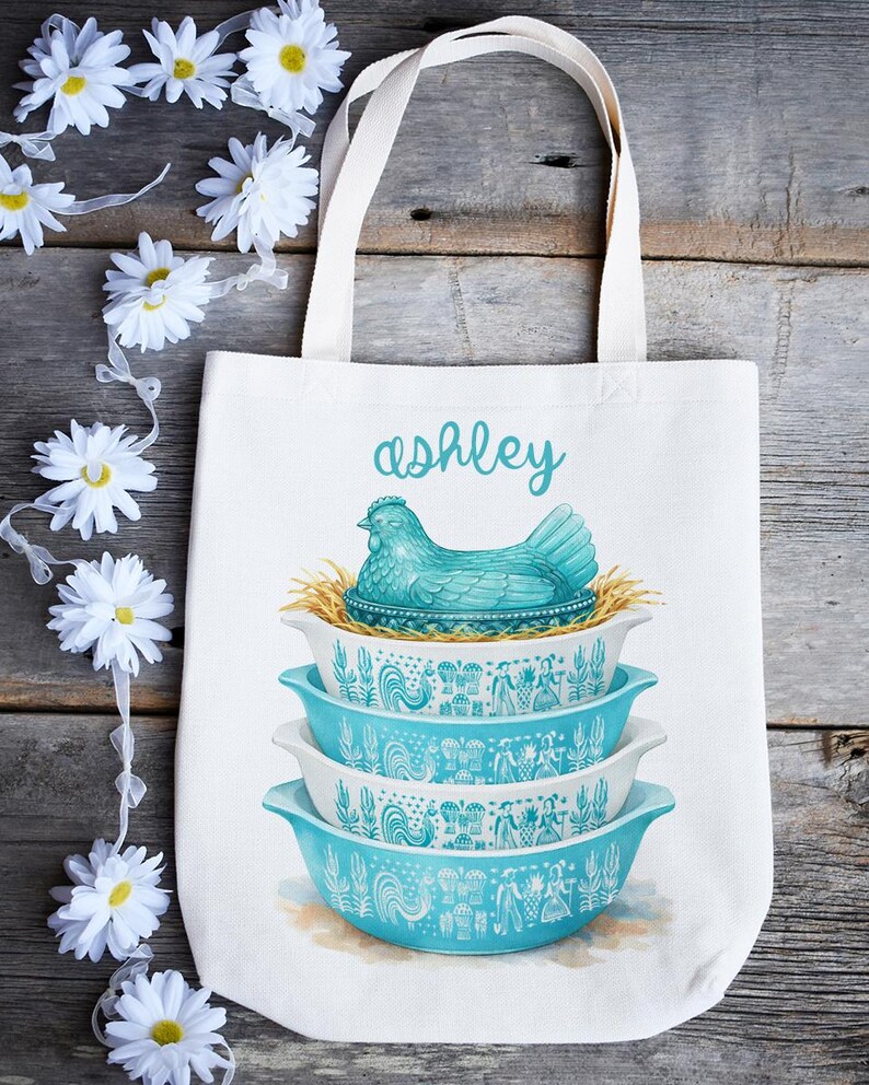May include: White tote bag featuring a stack of three turquoise and white bowls with a rooster and floral design. A turquoise hen sits atop the bowls, with the name "ashley" in script above. The bag has white handles.