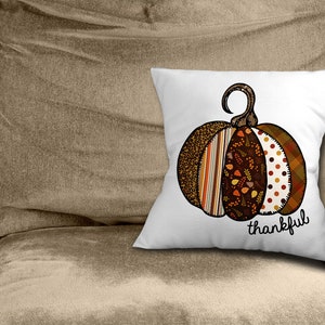 May include: White throw pillow featuring a brown, orange and white patchwork pumpkin design. The pumpkin has a stem and the text "thankful" is printed below it.