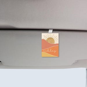 Op de afbeelding: Rechthoekige auto-geurverfrisser met een woestijnlandschapontwerp in tinten oranje, geel en beige. Het ontwerp bevat een zon met stralen en de naam "ashley" in een scriptlettertype. De geurverfrisser hangt aan een zonneklep in de auto.