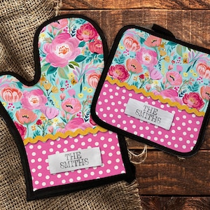 May include: A set of two pink and blue floral oven mitts with white polka dots and a personalized name tag that says "THE SMITHS".