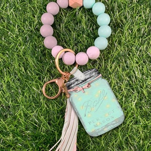 Op de afbeelding: Een lichtroze en lichtblauwe kralen sleutelhanger met een houten bedel en een mason jar bedel met de tekst "Ball Perfect Mason" en vuurvliegjes. De sleutelhanger heeft een kwast.