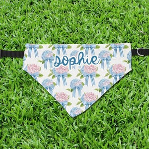 Puede incluir: Bandana para perros personalizada con un estampado floral de hortensias azules y rosas y lazos azules sobre un fondo crema. El nombre "Sophie" está escrito en azul. La bandana tiene un collar negro ajustable.