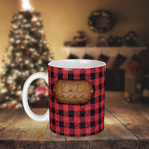 May include: A white ceramic mug with a red and black buffalo plaid design. The mug has a brown leather tag with the text "hot cocoa" on it.