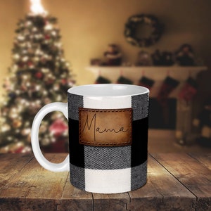 May include: White ceramic mug with a black and white buffalo plaid pattern. The mug has a brown leather patch with the word "Mama" written in cursive.