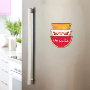 Może przedstawiać: Zestaw trzech drewnianych magnesów w kształcie miski w odcieniach złota, bieli i czerwieni. Środkowy magnes ma kwiatowy i ptasi wzór. Na dolnym magnesie znajduje się biały napis "the smiths". Magnesy są umieszczone na lodówce ze stali nierdzewnej.