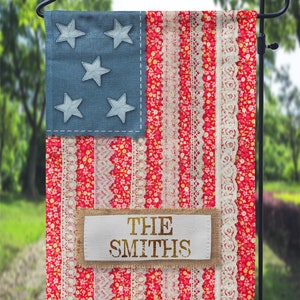 Op de afbeelding: Een rood, wit en blauwe tuin vlag met een wit stoffen label waarop "THE SMITHS" in gouden letters staat. De vlag heeft een patchwork ontwerp met kant en bloemenpatronen.
