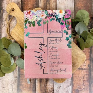 May include: A pink floral journal with a cross design and the name "Ashley" printed on it. The journal has inspirational quotes and bible verses printed on it, including "You are... Beautiful, Strong, Enough, Never alone, Always loved".