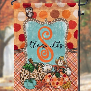 May include: A fall-themed garden flag with a blue and orange swirl design, the text "the smiths", and a variety of pumpkins, a scarecrow, and an owl. The background is a red and orange plaid pattern with white polka dots.