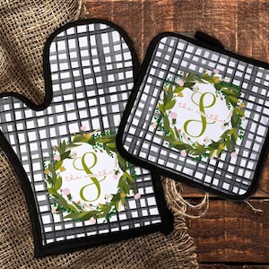 May include: A set of two black and white checkered oven mitts with a floral wreath design and the letter "S" in the center. The wreath has green leaves and pink flowers. The text "the smiths" is written in cursive below the letter.