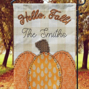 Puede incluir: Una bandera de jardín a cuadros beige y blanco con el texto "Hello, Fall! The Smiths" y una calabaza marrón, naranja y blanca con un tallo de arpillera.