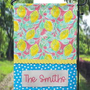 May include: A pink and green garden flag with a lemon pattern. The bottom of the flag is blue with white polka dots and a white rectangle with the text "The Smiths" in pink.