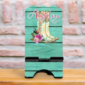 May include: A teal blue wooden phone stand with a rustic woodgrain design. The stand features a pair of brown cowboy boots with floral accents and the name "Ashley" in pink lettering.