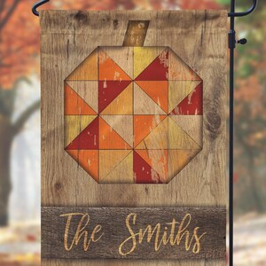 May include: A wooden garden flag featuring a geometric pumpkin design in shades of orange, red, and yellow. The flag has a rustic, distressed wood grain background and includes the text "The Smiths" in a cursive font.