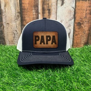 May include: A black and white trucker hat with a brown leather patch that says "PAPA" in white letters.