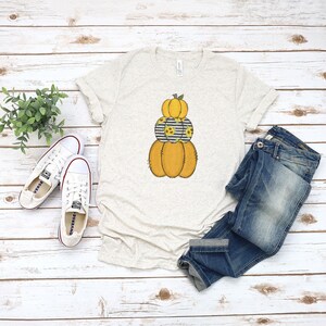 May include: A beige t-shirt with a stack of three pumpkins. The top pumpkin is orange with a green stem. The middle pumpkin is black and white striped with sunflowers. The bottom pumpkin is orange. The t-shirt is laying on a white wooden surface with a pair of white sneakers and blue jeans.