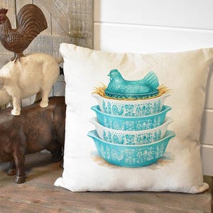 May include: A decorative pillow featuring a stack of four turquoise and white patterned bowls, topped with a turquoise hen on a nest. The pillow is on a wooden bench with farm animal figurines.