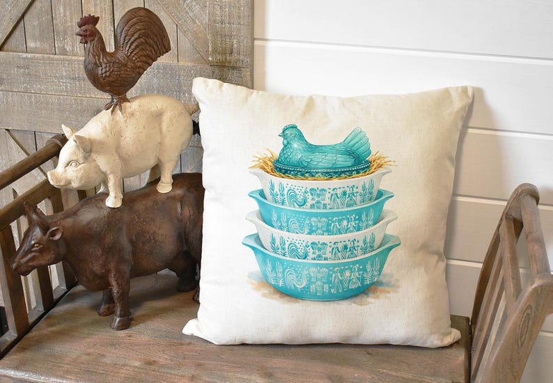 May include: A decorative pillow featuring a stack of four turquoise and white patterned bowls, topped with a turquoise hen on a nest. The pillow is on a wooden bench with farm animal figurines.