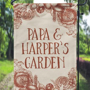 以下が含まれることがあります： ベージュの背景と茶色の野菜の縁取りのあるガーデンフラッグ。"Papa & Harper's Garden"という文字が茶色で印刷されています。