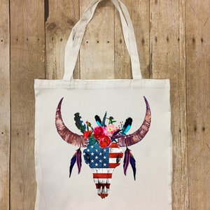 May include: A white tote bag with a colorful bull skull design. The skull is decorated with flowers and feathers, and the skull is painted with the American flag.