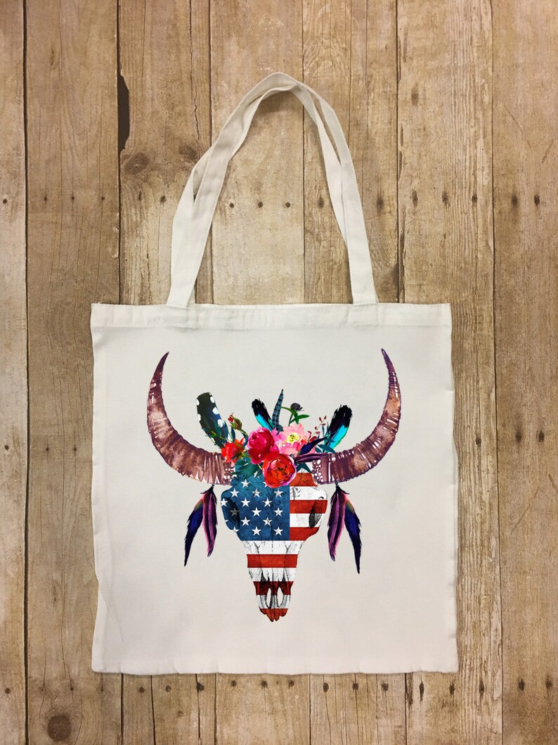 May include: A white tote bag with a colorful bull skull design. The skull is decorated with flowers and feathers, and the skull is painted with the American flag.