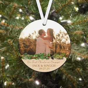 Könnte beinhalten: Runder Weihnachtsschmuck mit einem Foto eines Paares, das sich umarmt. Der Schmuck hat ein weißes Band zum Aufhängen und den Text "Our First Christmas Engaged" mit Namen und Datum. Grün und rote Beeren schmücken den unteren Rand.