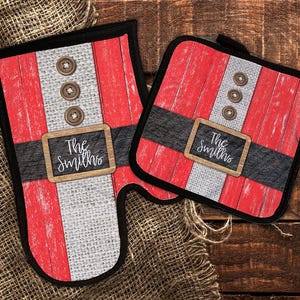 Puede incluir: Dos agarraderas de olla rojas y blancas con un cinturón negro y un letrero de pizarra que dice "The Smiths". Las agarraderas de olla tienen forma de traje de Papá Noel.