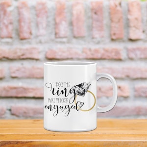 May include: White ceramic mug with a black and gold design. The mug features a diamond ring and the text "Does this ring make me look engaged?"