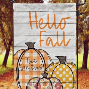 May include: A white wooden sign with the text "Hello Fall" in orange. Three pumpkins are stacked on the sign, the top pumpkin is yellow with white polka dots, the middle pumpkin is orange with white and orange checkered pattern and the text "The Smiths" in black, and the bottom pumpkin is a floral pattern with orange, yellow, and pink flowers.