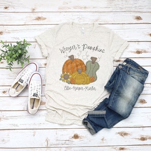 Pode incluir: Uma camiseta bege claro com o texto "Weezer's Pumpkins" e "Ellie-Harper-Karter" impresso nela. A camiseta apresenta um gráfico de três abóboras com um girassol.