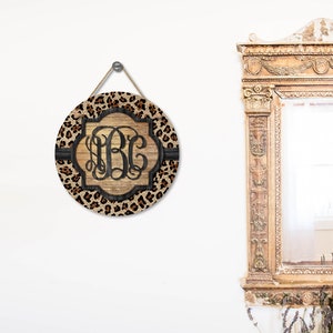 Op de afbeelding: Een rond houten bord met een luipaardprint achtergrond en een zwart monogram in het midden. Het bord hangt aan een touwtje en heeft een rustieke houtnerf.