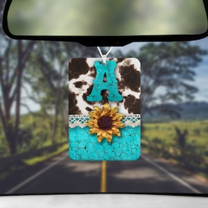 May include: A turquoise blue car air freshener with a cow print background, a white lace trim, a sunflower, and the letter "A" in turquoise blue.