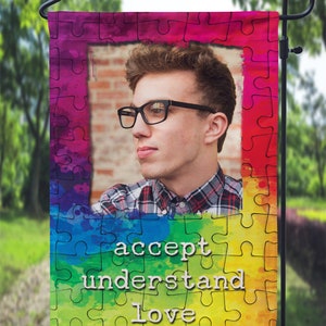 May include: A garden flag with a rainbow watercolor background and a puzzle piece pattern. The flag features a photo of a person with glasses and the words "accept understand love".