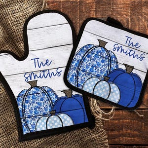 May include: A set of two blue and white patterned oven mitts with the text "The Smiths" on each mitt. The mitts feature a woodgrain background and a design of pumpkins.