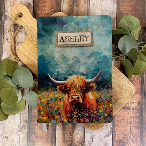 Puede incluir: Una cubierta de cuaderno personalizada con una pintura de acuarela de una vaca de las Tierras Altas en un campo de flores silvestres. El nombre "ASHLEY" está impreso en una etiqueta de arpillera.