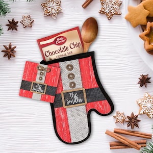 Puede incluir: Manopla de horno roja y blanca con forma de Papá Noel con un cinturón negro y una etiqueta de pizarra que dice "The Smiths". La manopla está llena de una caja de mezcla para galletas con chispas de chocolate Betty Crocker.