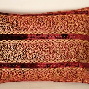 May include: A red velvet cushion with gold embroidered geometric patterns. The cushion is rectangular in shape and features a repeating design of diamonds and floral motifs.