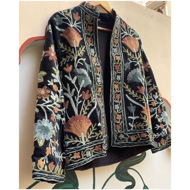 May include: Black embroidered jacket with a mandarin collar and open front. The jacket is adorned with colourful floral embroidery in shades of orange, green, and blue. It is displayed on a wooden hanger.