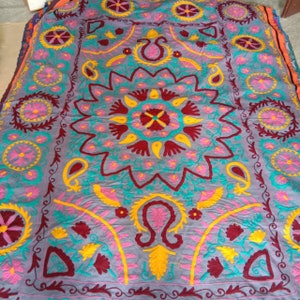 May include: A gray fabric rug with intricate embroidery in shades of pink, yellow, blue, and maroon. The design features a large central floral motif surrounded by smaller floral and paisley patterns.