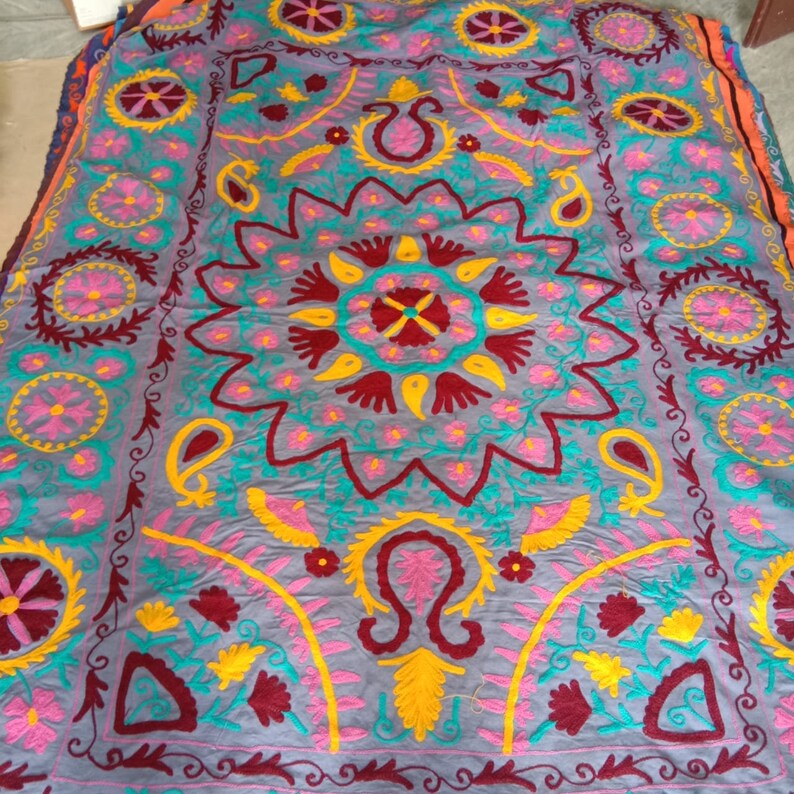 May include: A gray fabric rug with intricate embroidery in shades of pink, yellow, blue, and maroon. The design features a large central floral motif surrounded by smaller floral and paisley patterns.