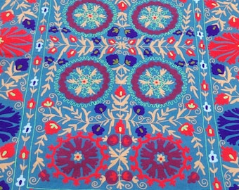 May include: A blue embroidered fabric with a floral pattern in shades of red, pink, and blue. The fabric has a square shape and is decorated with a central motif of four circles.