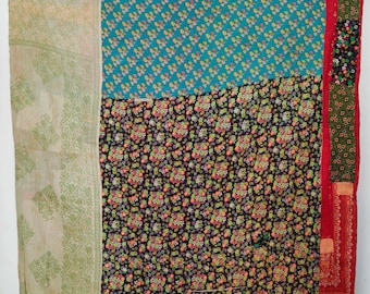 Op de afbeelding: Een rechthoekige patchwork quilt met een groot blauwgroen gedeelte met een klein bloemenpatroon, een zwart gedeelte met een dichte veelkleurige bloemenprint, een lichtgroen gedeelte met een subtiel bladmotief en een rode verticale rand.