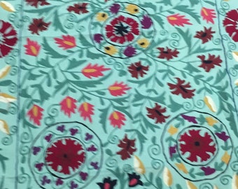 May include: A teal fabric with a floral embroidery pattern. The flowers are red, pink, and yellow with green leaves. The fabric has a white border.