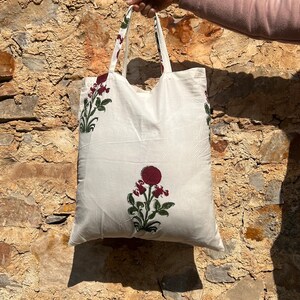 May include: White cotton tote bag with red and green floral print. The bag has two handles and is held by a hand.